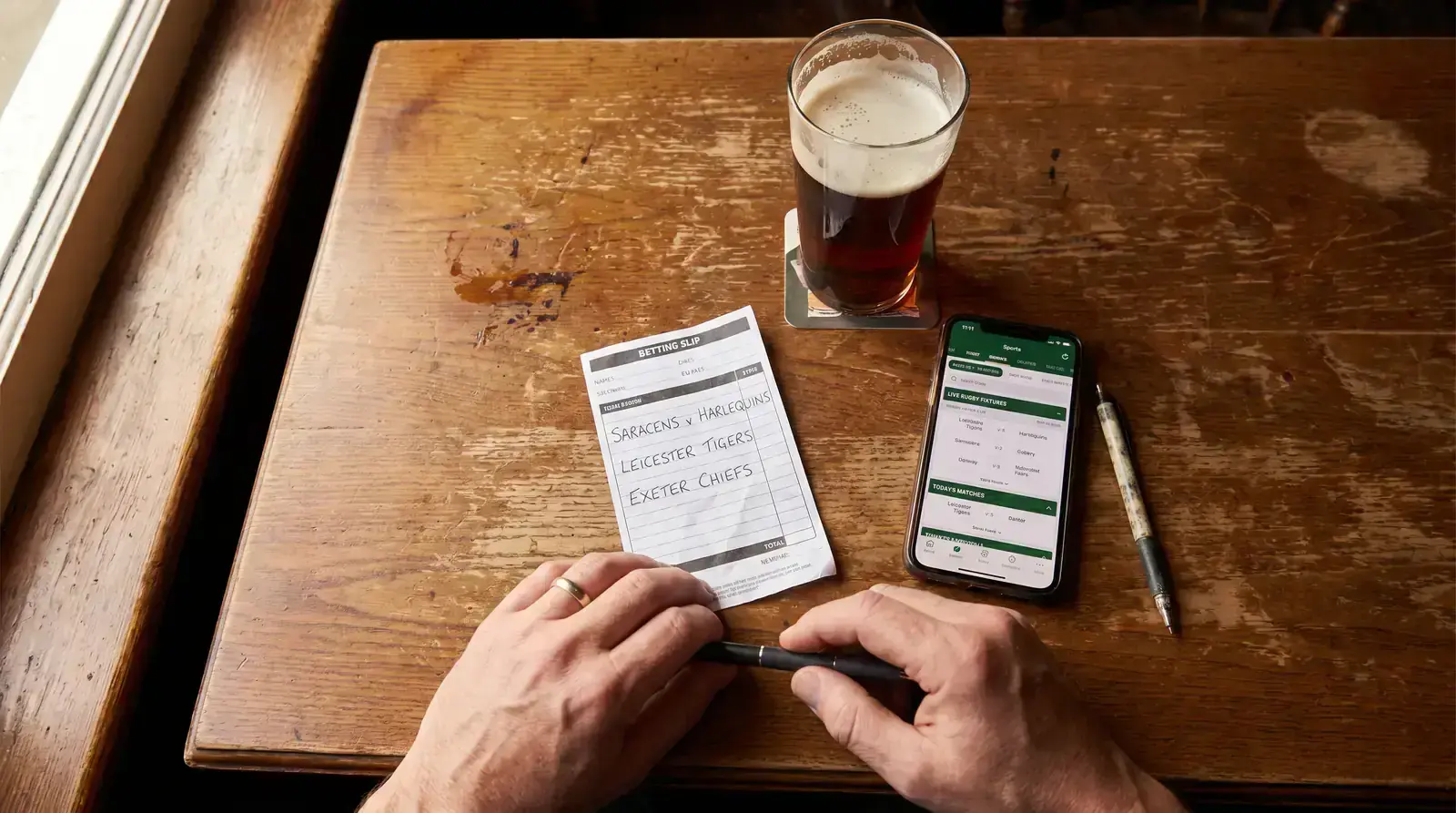 Hands holding a paper rugby betting slip next to a phone showing match markets